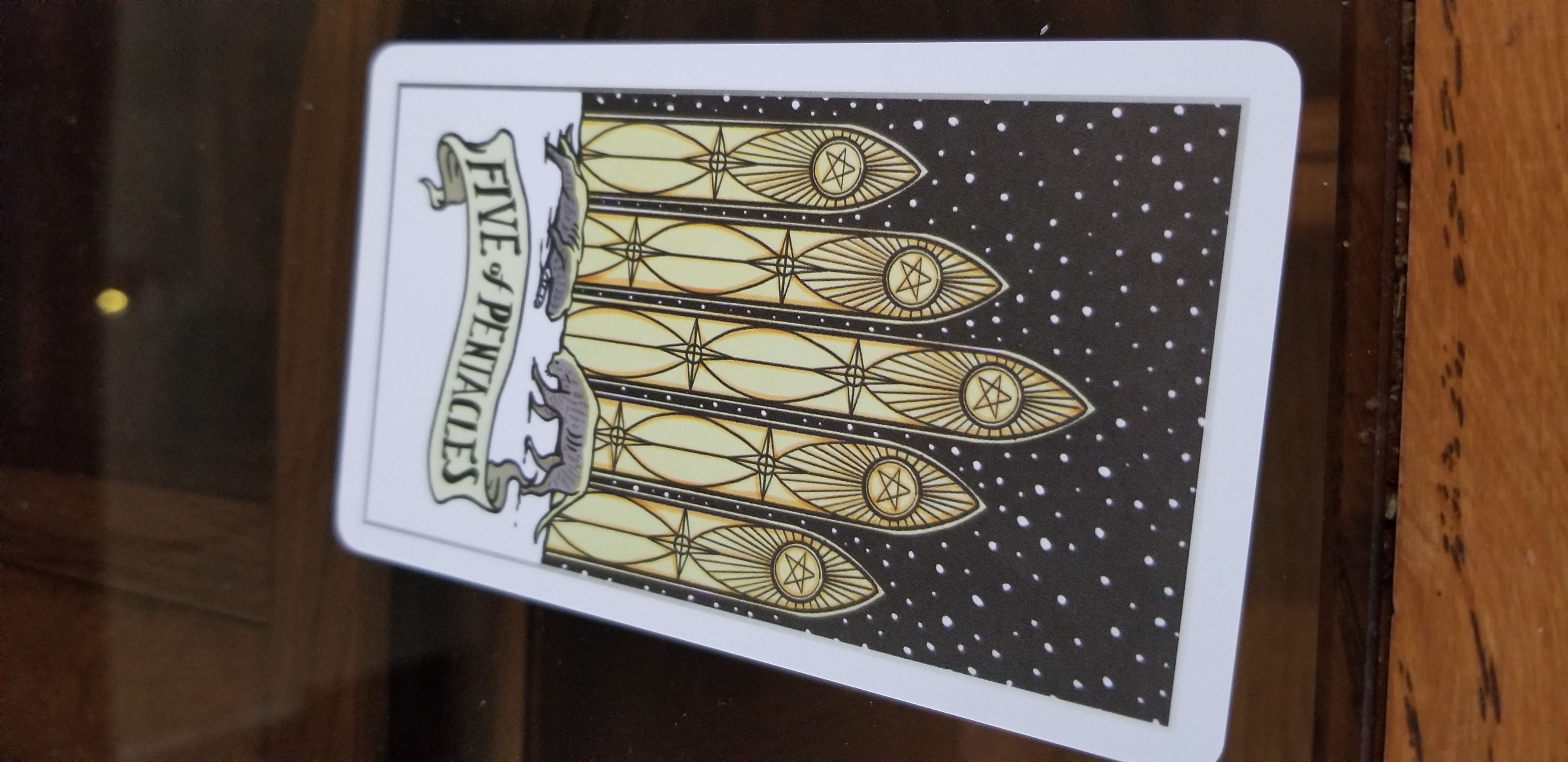 an inverted five of pentacles from the stray cat tarot deck. the five of discs features a cat with a bandaged paw being approached by another cat. behind them are five tall gothic windows, presumably a cathedral, bearing a pentacle upon each.