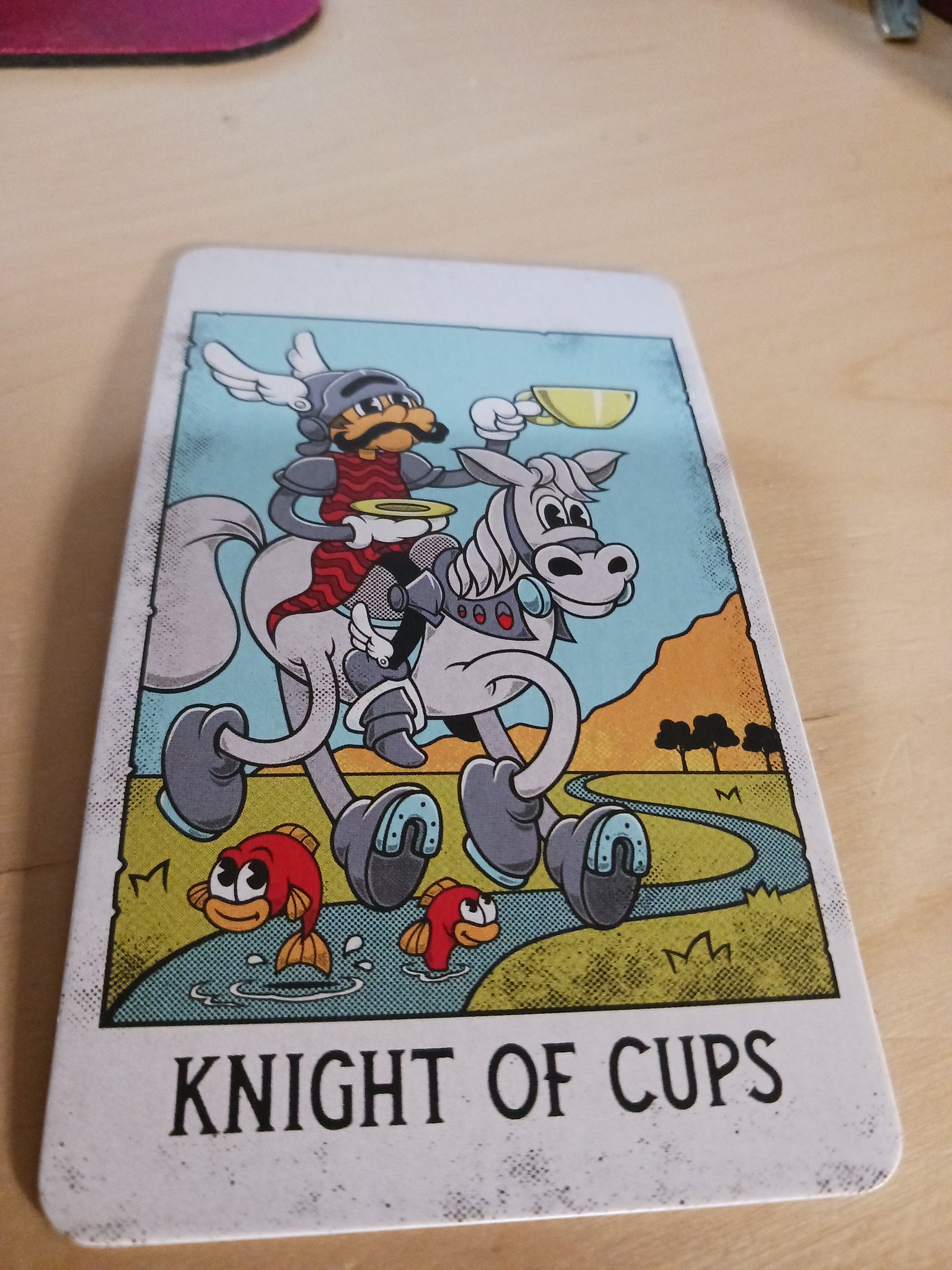 an upright knight of cups from the mystical medleys deck. the image is a knight astride his horse while crossing a stream. he is holding a cup aloft. two cartoon fish are jumping out of the water underneath the steed.